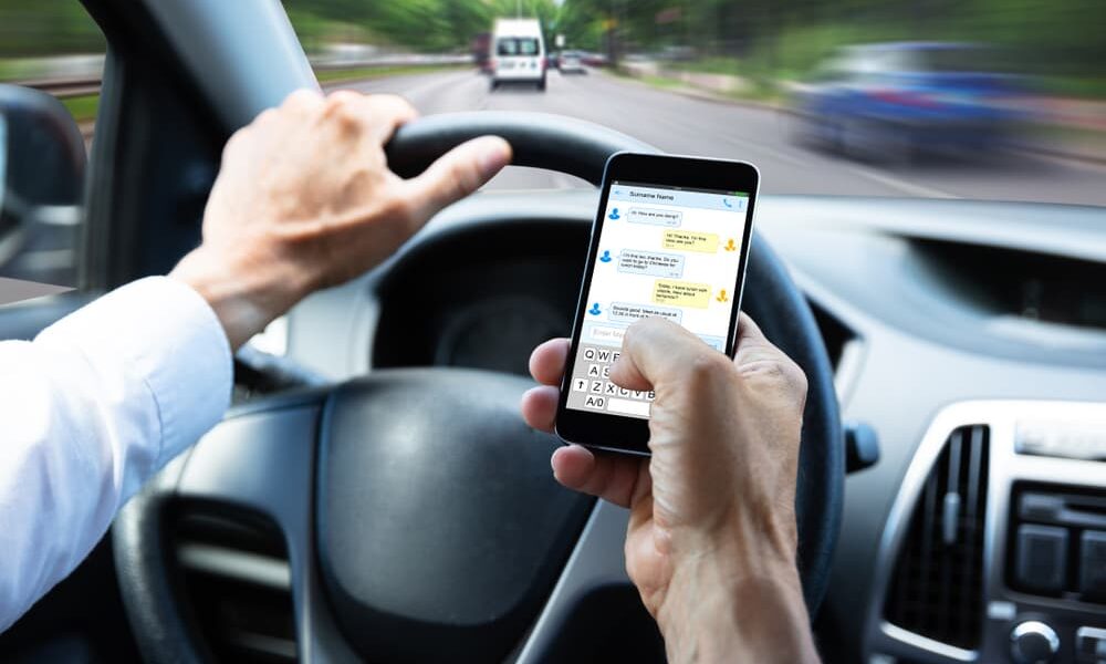 A close-up of a man's hand typing a text message on a mobile phone while driving a car.