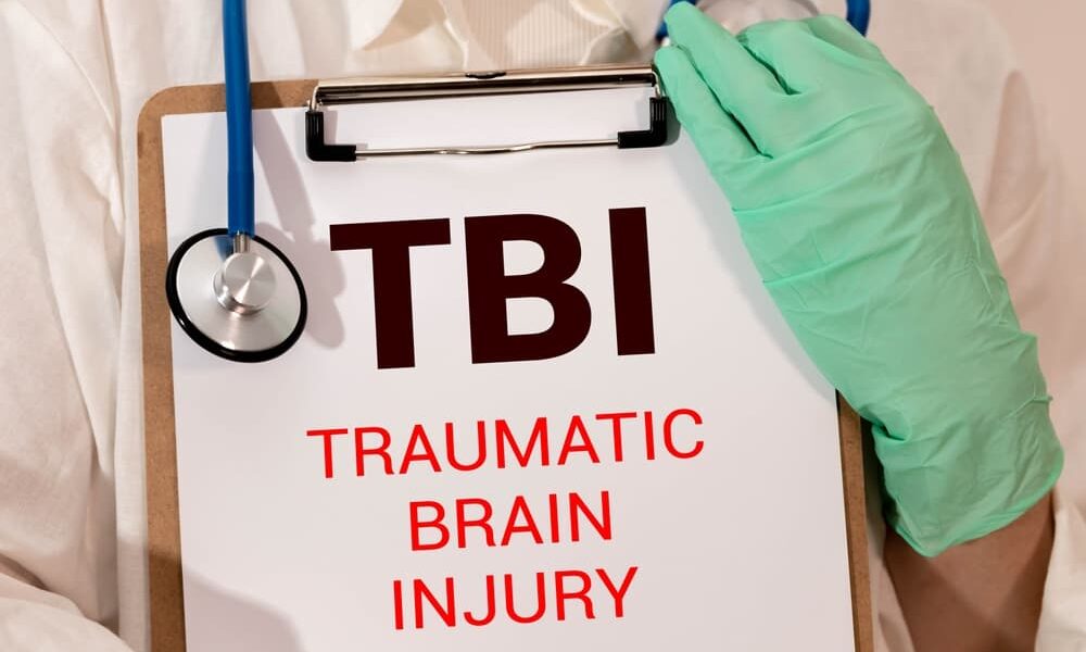 "TBI Traumatic Brain Injury" written on a white note, set against a background of dollar bills and a pen. A visual representation of medical and financial aspects related to TBIs.