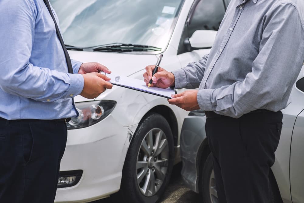 An insurance agent examines a damaged car while the customer signs a claim report form following a traffic accident, illustrating the insurance claim process.







