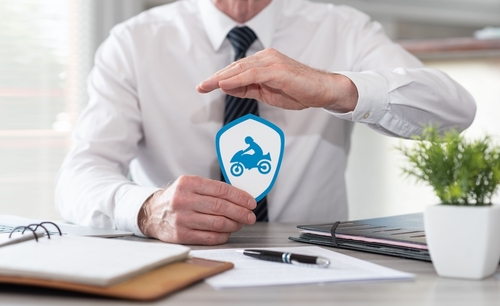 A man in formal attire sitting at a desk, holding a shield-shaped icon with a motorcycle symbol, symbolizing motorcycle insurance or legal protection.