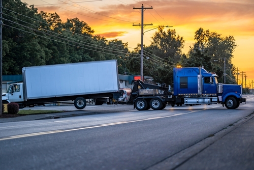 A blue semi-truck towing a damaged white box truck on a street at sunset, suggesting an accident recovery scene.